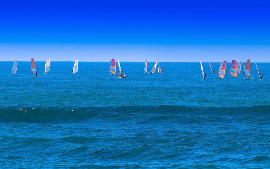 A fleet of windsurfers with brightly coloured sails on a vivid blue sea