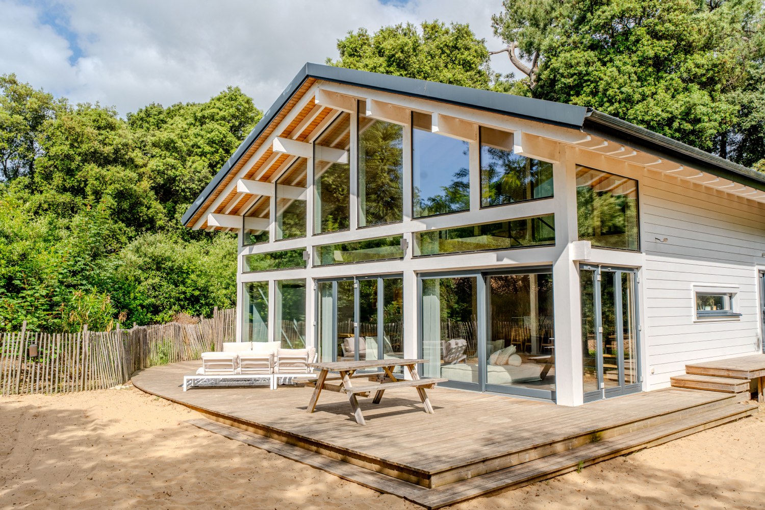Le Chalet Moana vu du jardin : façade en bois massif, grandes baies vitrées, terrasse et table de pique-nique sous un ciel bleu