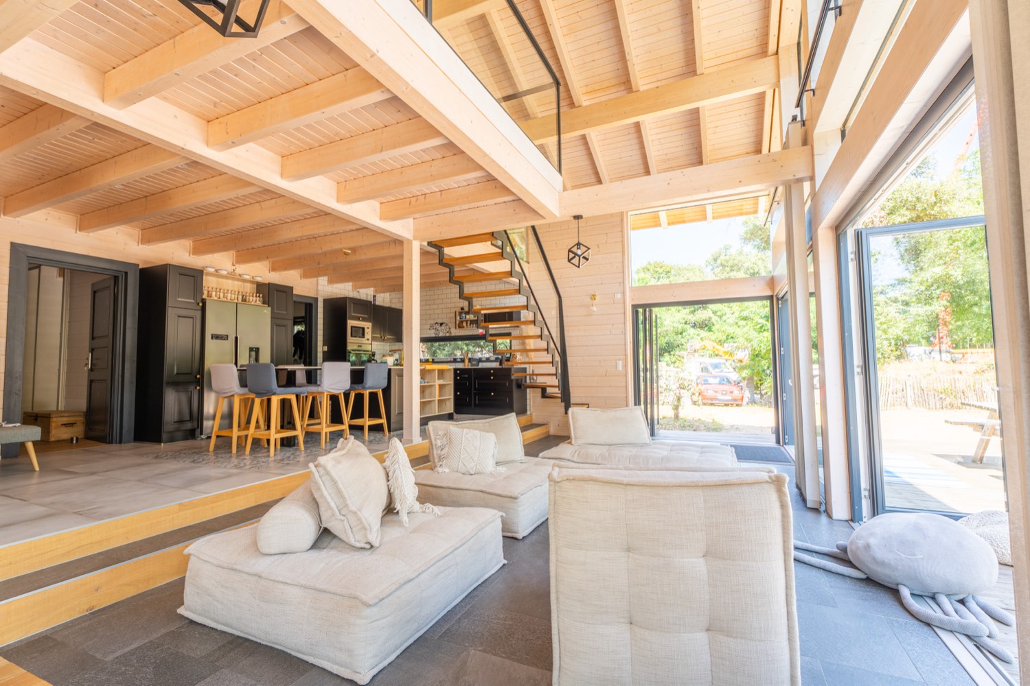 Grande pièce de vie ouverte du Chalet Moana avec salon en lin clair, cuisine noire, escalier et mezzanine sous charpente en bois clair