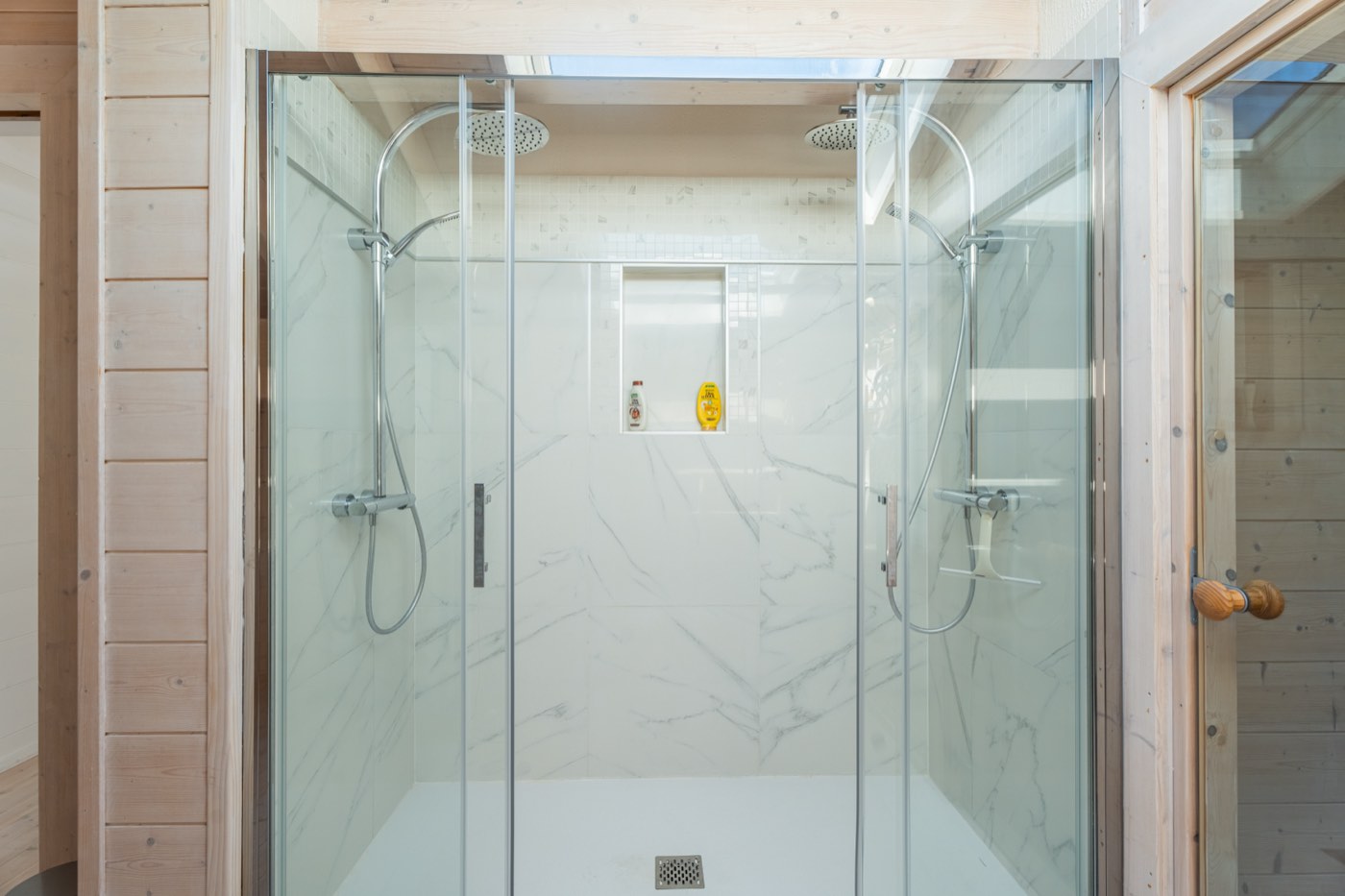 Grande double douche à l'étage du Chalet Moana : deux colonnes avec pomme de pluie, parois vitrées, faïence effet marbre et ciel de toit au-dessus