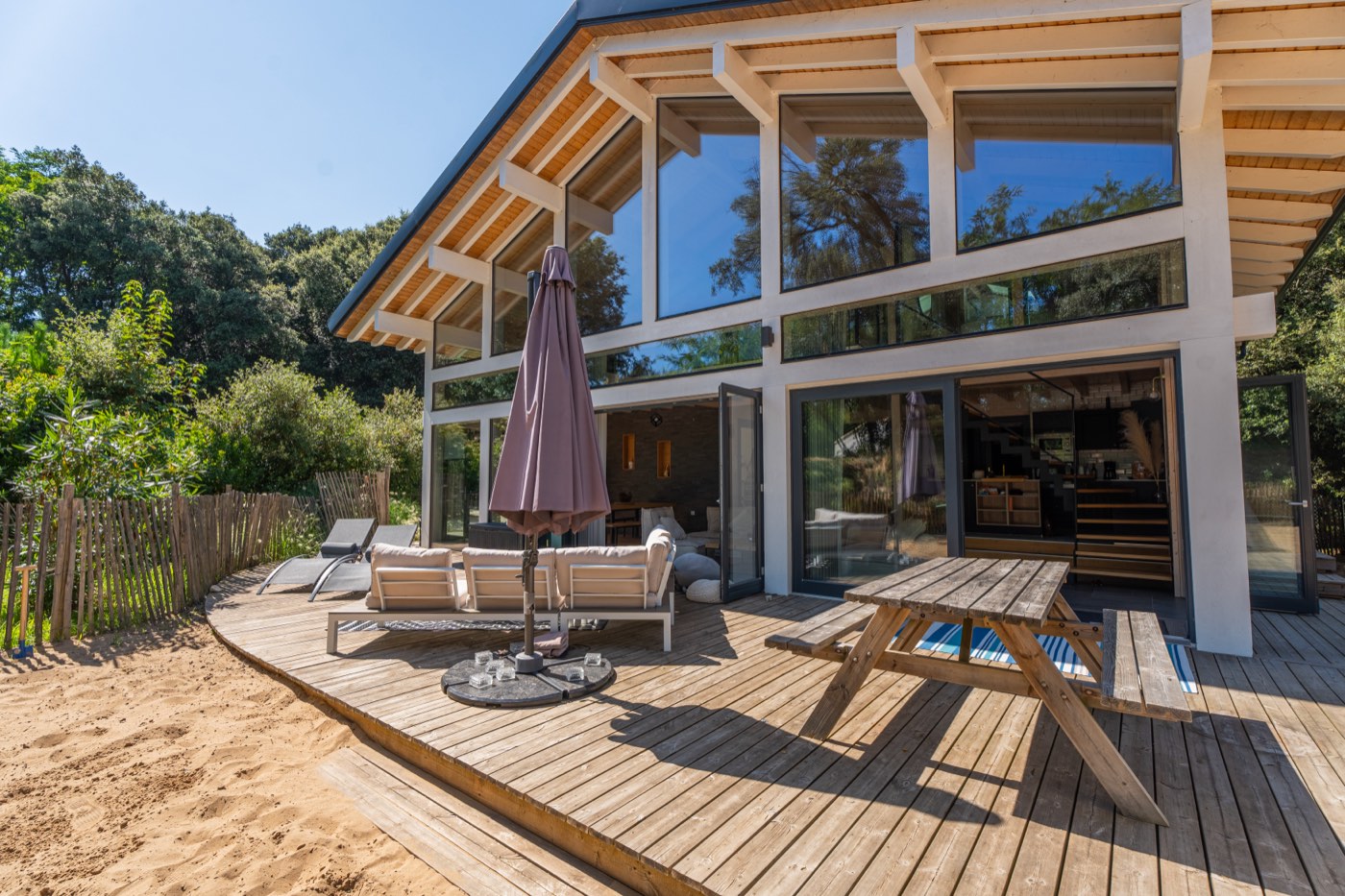 Terrasse en bois du Chalet Moana avec bains de soleil, parasol et table de pique-nique, le sable commençant au pied du plancher